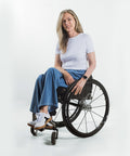 A woman with blonde hair sits smiling in a black manual wheelchair, wearing a white t-shirt, blue wide-leg jeans, and white sneakers against a clean white studio background.