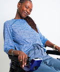A smiling woman with long dark braids sits in a wheelchair wearing a heathered blue long-sleeve top and matching blue pants against a plain light background.
