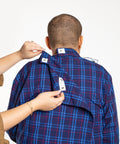 A man viewed from behind wearing a blue and red plaid button-down shirt, while another person's hands adjust the collar and open the back flap of the shirt against a plain white background.
