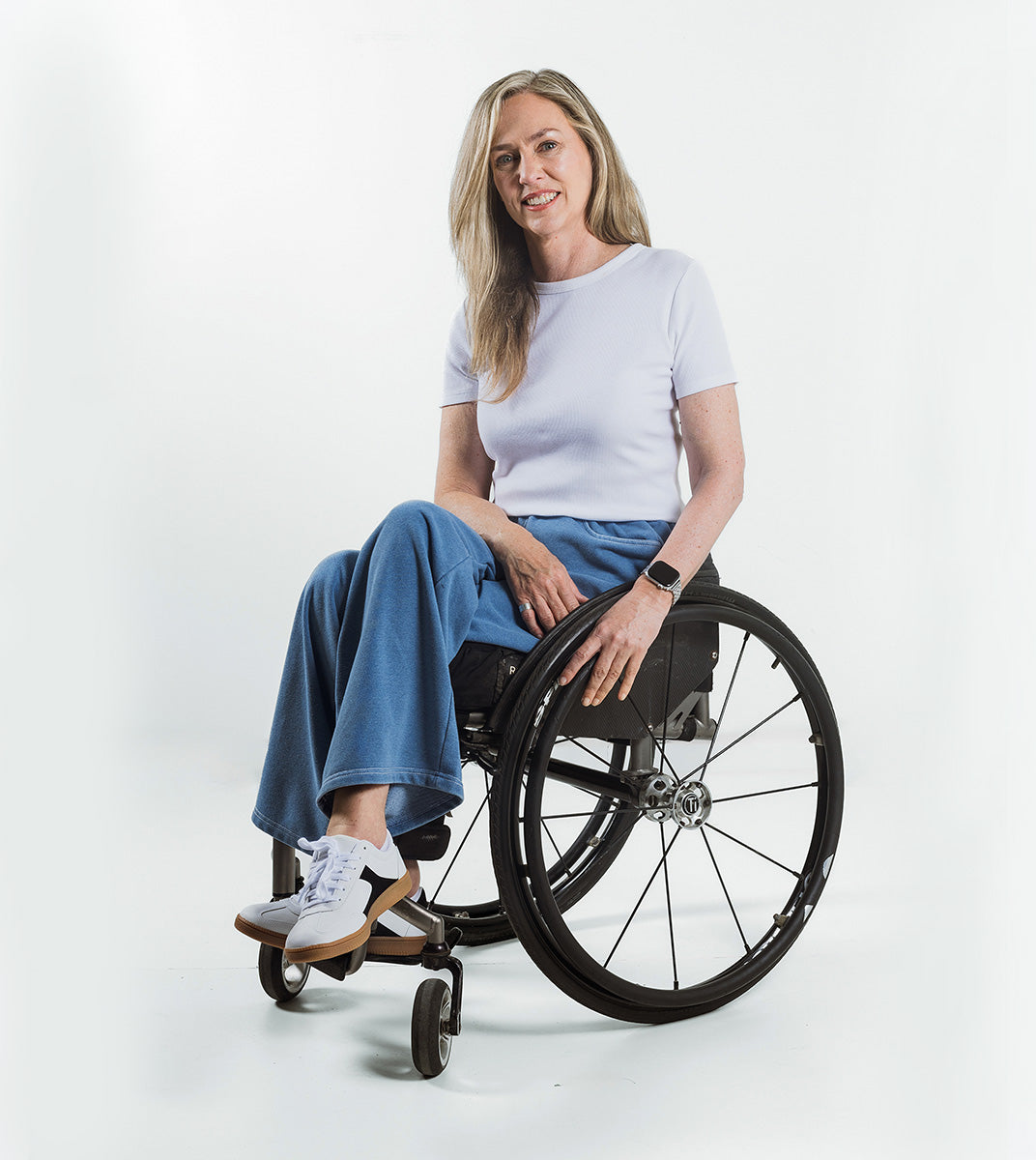 A woman with blonde hair sits smiling in a black manual wheelchair, wearing a white t-shirt, blue wide-leg jeans, and white sneakers against a clean white studio background.