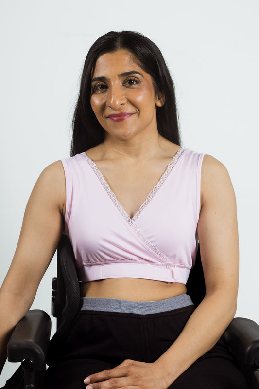 A woman with long dark hair sits in a black armchair, wearing a light pink wrap-style crop top with lace trim and black pants against a plain light gray background.