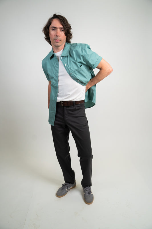 A man posing in a studio setting, wearing a teal short-sleeve button-up shirt over a white t-shirt, black trousers, and gray sneakers.