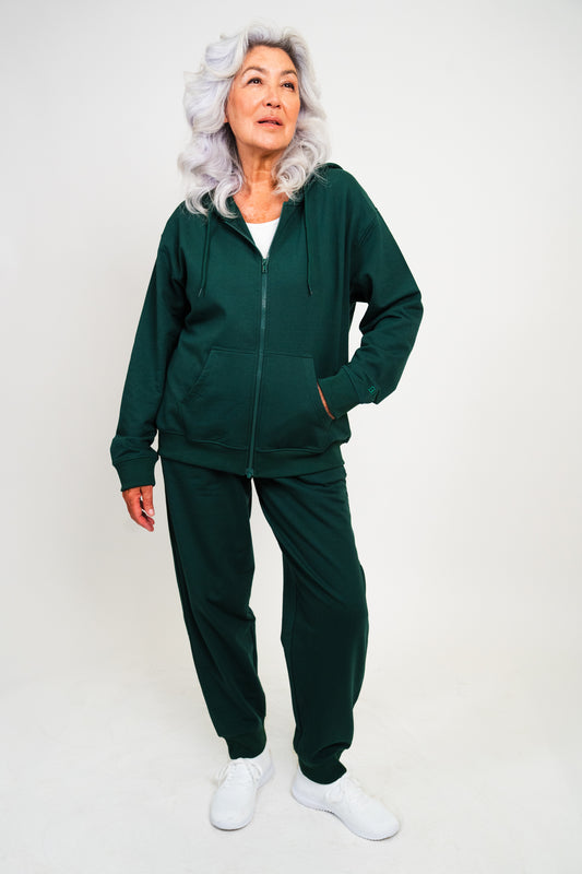 An older woman with silver-gray hair models a dark green zip-up hoodie and matching sweatpants set, paired with white sneakers, against a plain light gray studio background.