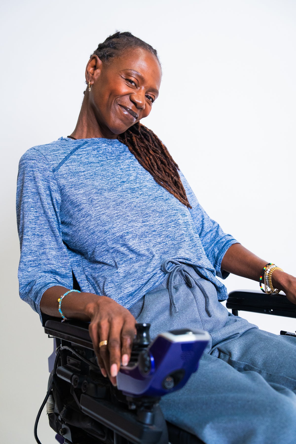 A smiling woman with long dark braids sits in a wheelchair wearing a heathered blue long-sleeve top and matching blue pants against a plain light background.