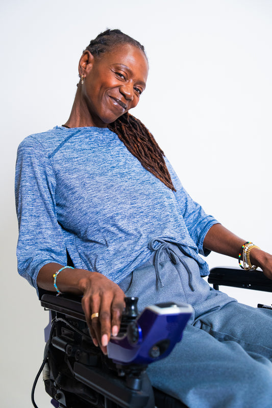 A smiling woman with long dark braids sits in a wheelchair wearing a heathered blue long-sleeve top and matching blue pants against a plain light background.