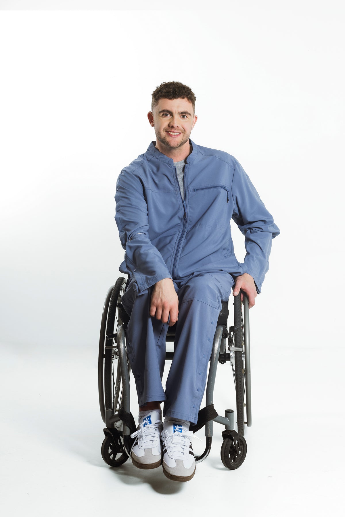 A man wearing a blue scrub set sits in a wheelchair against a plain white studio background.