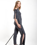 A woman modeling a dark gray off-the-shoulder top and matching slim-fit pants with side slits, styled with black slip-on shoes and holding a black cane against a plain white studio background.