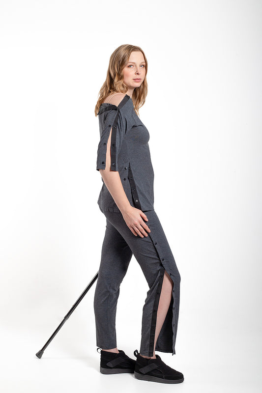 A woman modeling a dark gray off-the-shoulder top and matching slim-fit pants with side slits, styled with black slip-on shoes and holding a black cane against a plain white studio background.